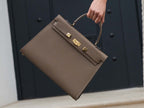 A large taupe colored Italian calf leather handbag with pebble print texture, gold plated hardware, and a top handle with a detachable strap held by a hand against a dark background