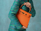 Woman holding a large orange, natural grainy print Italian calf leather handbag with top handle, detachable strap and gold plated hardware against a light blue background