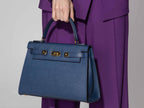 A large navy Italian leather, natural print handbag with gold hardware, a top handle with a detachable strap on a white background held by a person wearing a purple outfit on a gray background