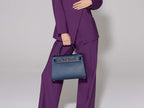 Woman wearing a purple suit holding a large navy Italian leather, natural print handbag with gold hardware, a top handle with a detachable strap against a plain background