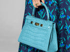 A large turquoise , ‘croc’ print Italian calf leather handbag with top handle, detachable strap and gold plated hardware held by a person wearing a dark blue floral dress.