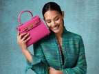 Woman holding a bright pink, medium-sized, grainy print Italian calf leather handbag with gold-plated hardware, detachable strap and a top handle against a blue background