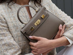 Woman holding a taupe colored, medium-sized handbag made from grainy print Italian calf leather, featuring gold-plated hardware and a top handle with a detachable shoulder strap.