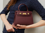Woman holding a burgundy wine coloured, medium-sized Italian leather handbag with a pebble print and gold plated hardware, featuring a top handle and a detachable shoulder strap. against a neutral background