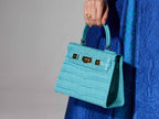 A turquoise 'Croc' print Italian calf leather handbag with gold plated hardware and a top handle held by a person wearing a blue dress against a gray background