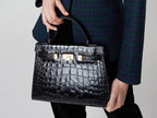 Woman holding a large black, ‘croc’ print Italian calf leather handbag with top handle, detachable strap and gold plated hardware.