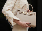 Woman carrying a large beige, natural grainy print Italian calf leather handbag with top handle, detachable strap and gold plated hardware.