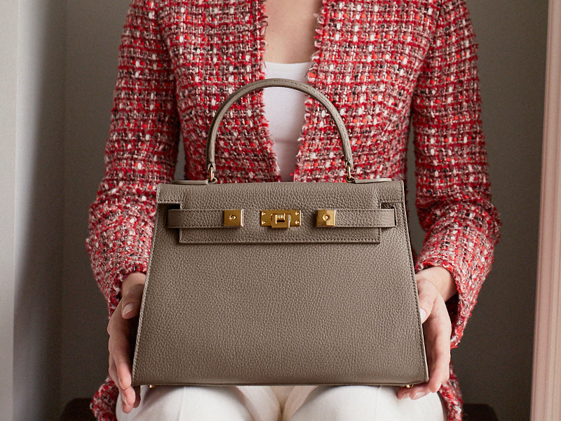 Woman holding a large taupe coloured, natural grainy print Italian calf leather handbag with top handle, detachable strap and gold plated hardware.