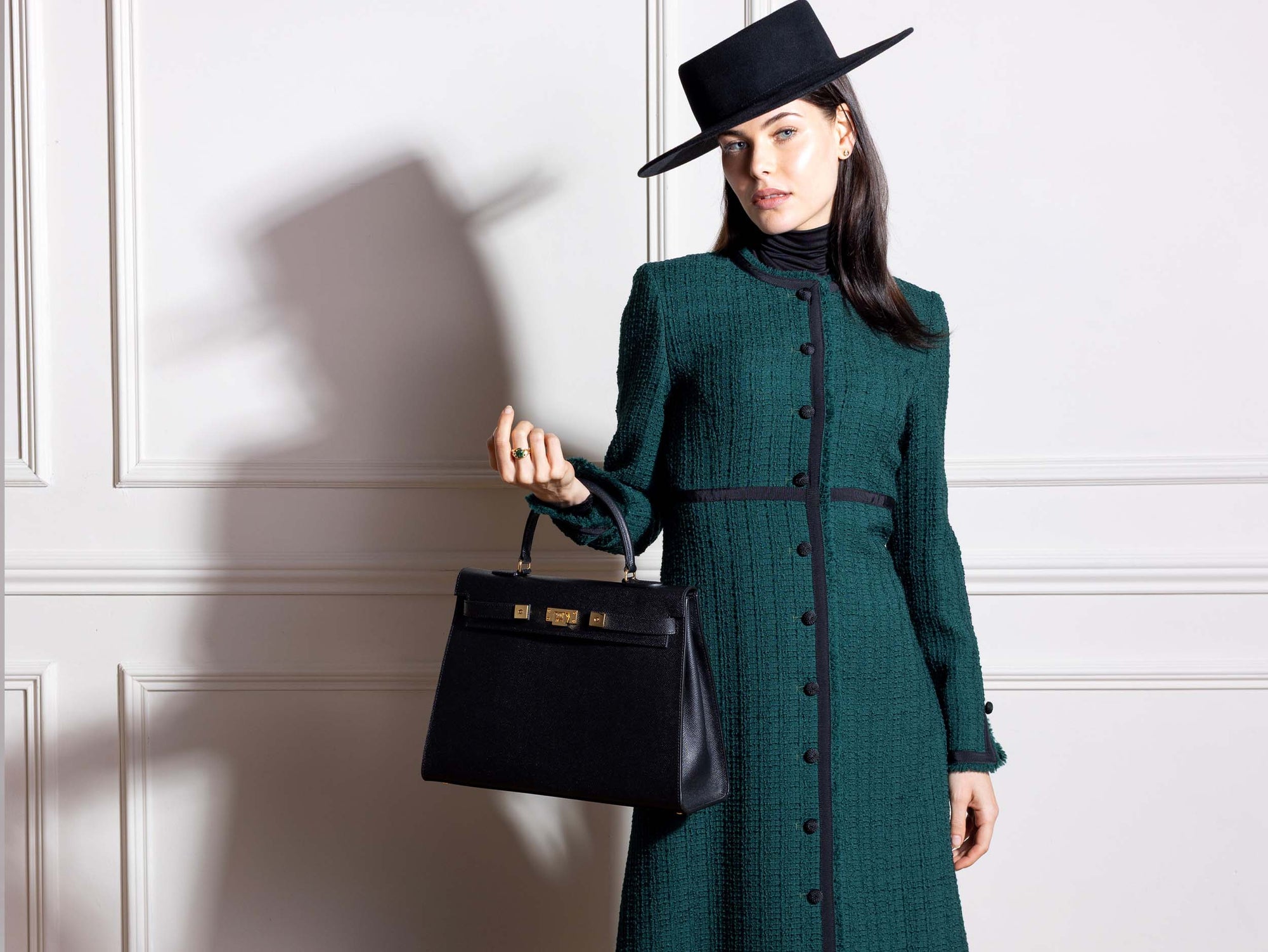Woman in a green coat and black hat holding a black Maya Grande Dolomite handbag with gold plated hardware and a pebble print texture, featuring a top handle, detachable strap and gold accents against a white wall.