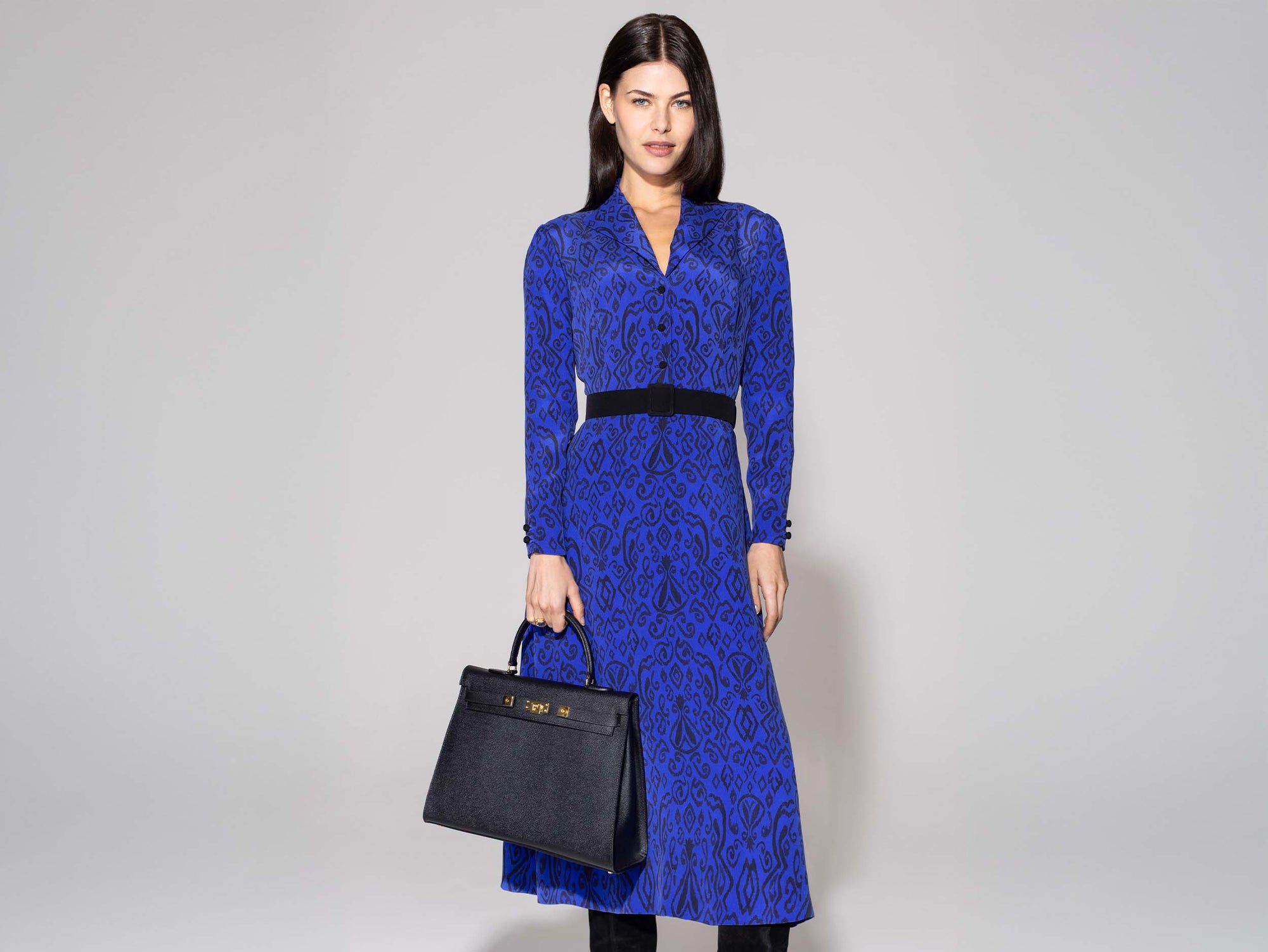 Woman wearing a blue lace dress holding a black Maya Grande Dolomite handbag with gold plated hardware and a pebble print texture, featuring a top handle, detachable strap and gold accents on a gray background