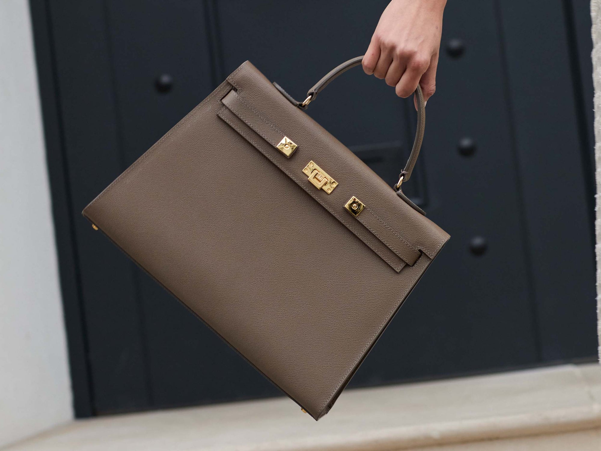 A large taupe colored Italian calf leather handbag with pebble print texture, gold plated hardware, and a top handle with a detachable strap held by a hand against a dark background