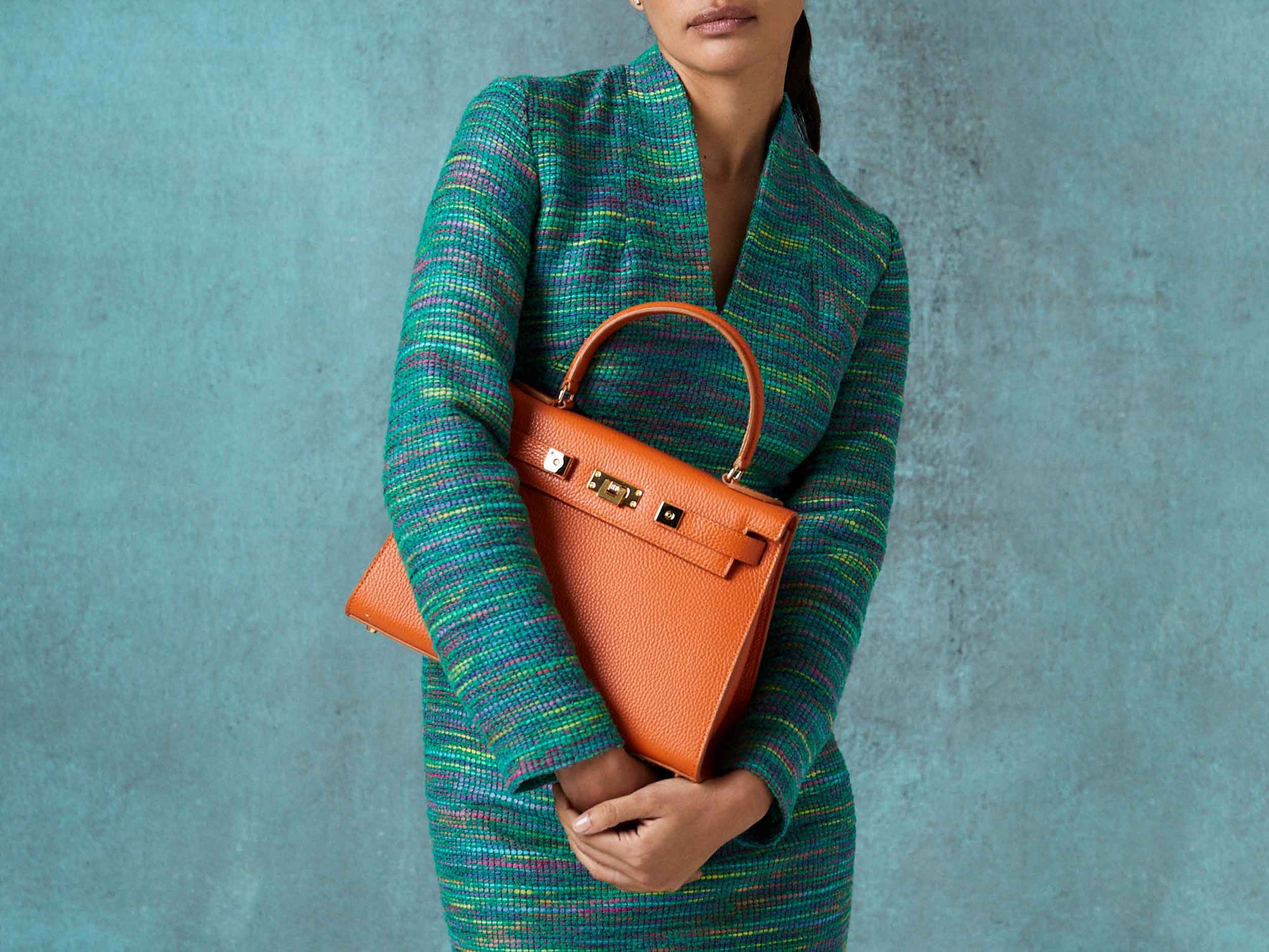 Woman holding a large orange, natural grainy print Italian calf leather handbag with top handle, detachable strap and gold plated hardware against a textured blue background