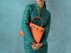 Woman holding a large orange, natural grainy print Italian calf leather handbag with top handle, detachable strap and gold plated hardware against a textured blue background