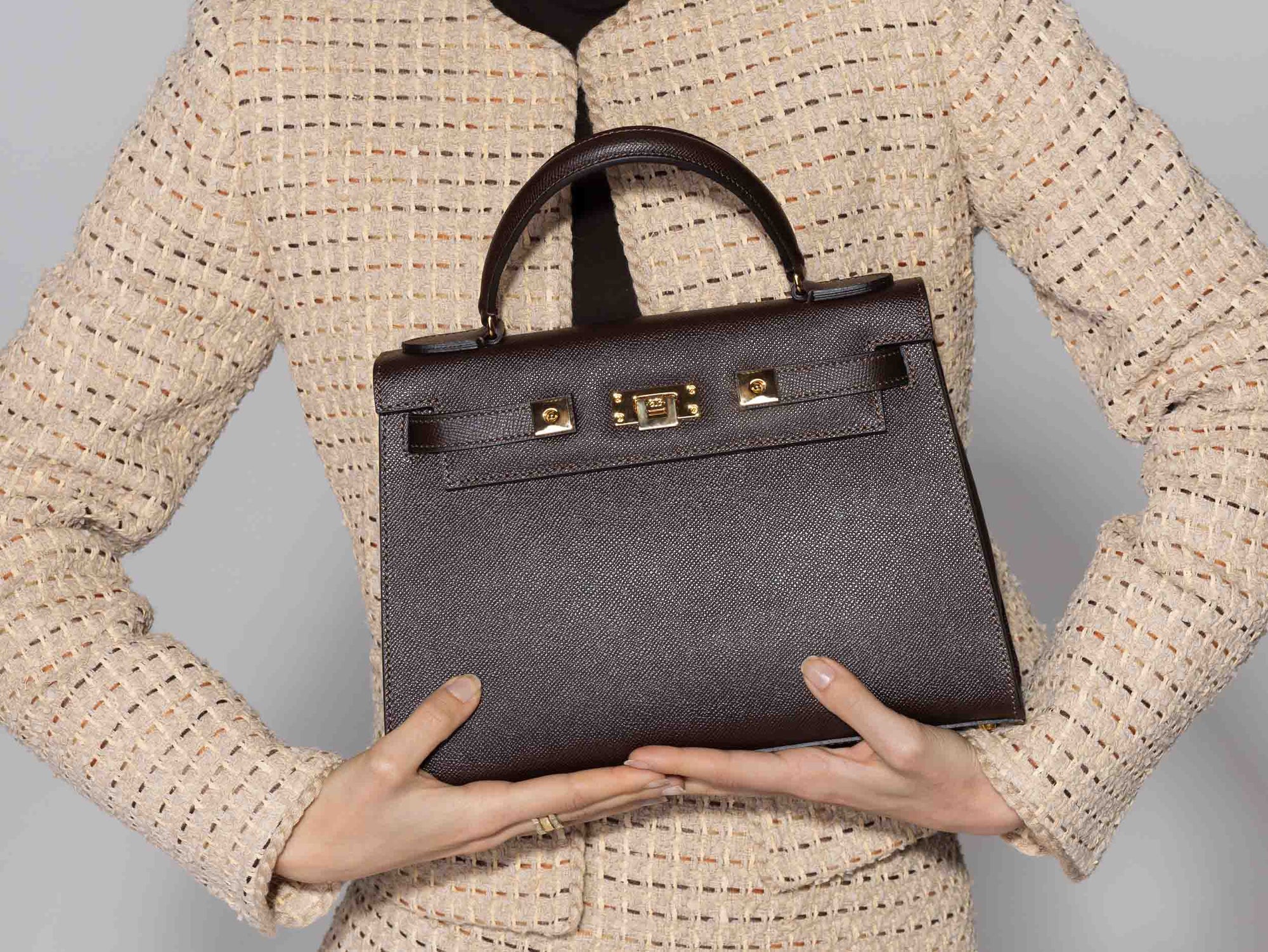 Person holding a large brown Italian leather, natural print handbag with gold hardware, a top handle with a detachable strap on a white background against a neutral background