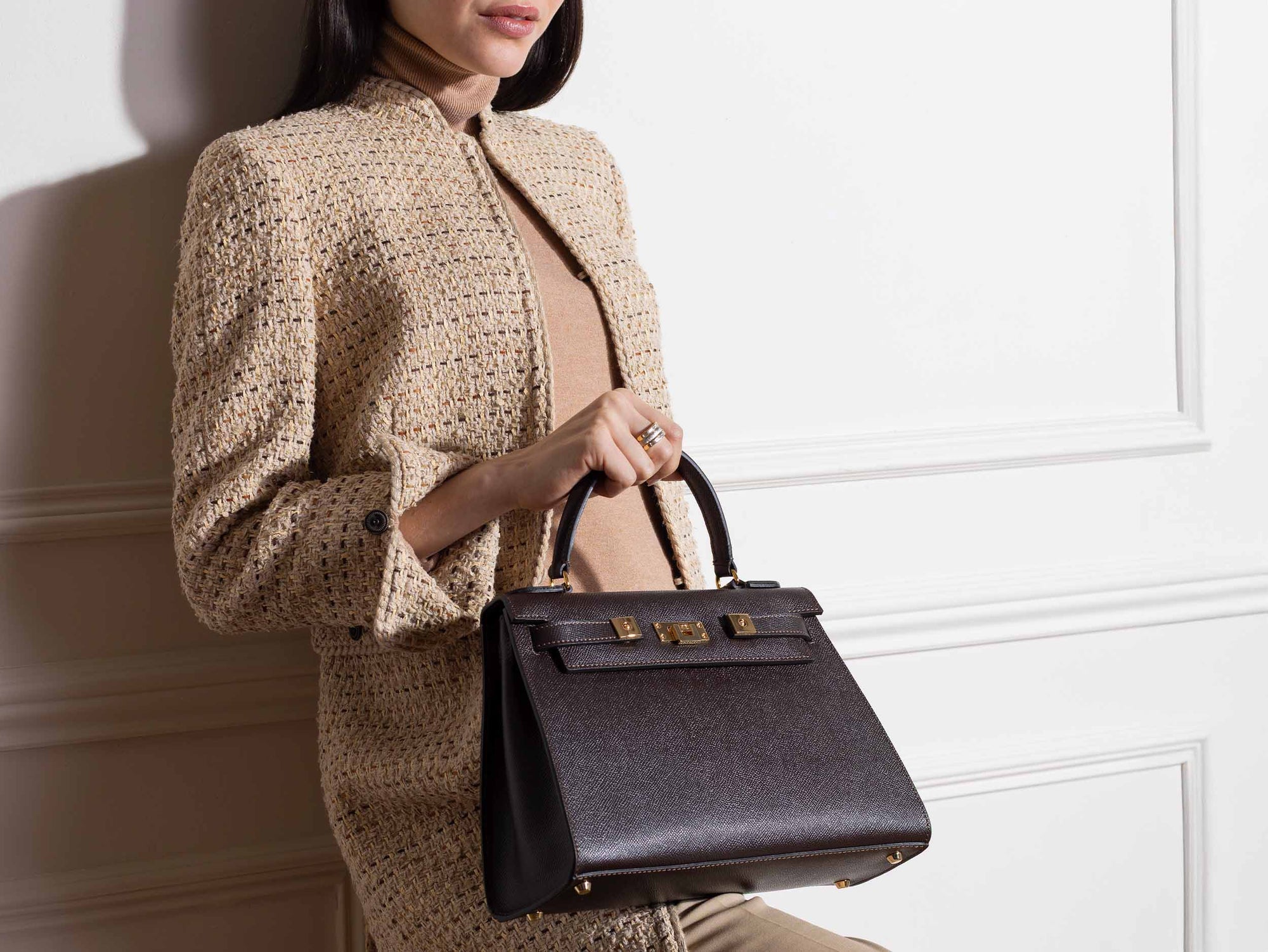 Woman holding a large brown Italian leather, natural print handbag with gold hardware, a top handle with a detachable strap on a white background against a white wall