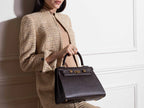 Woman holding a large brown Italian leather, natural print handbag with gold hardware, a top handle with a detachable strap on a white background against a white wall