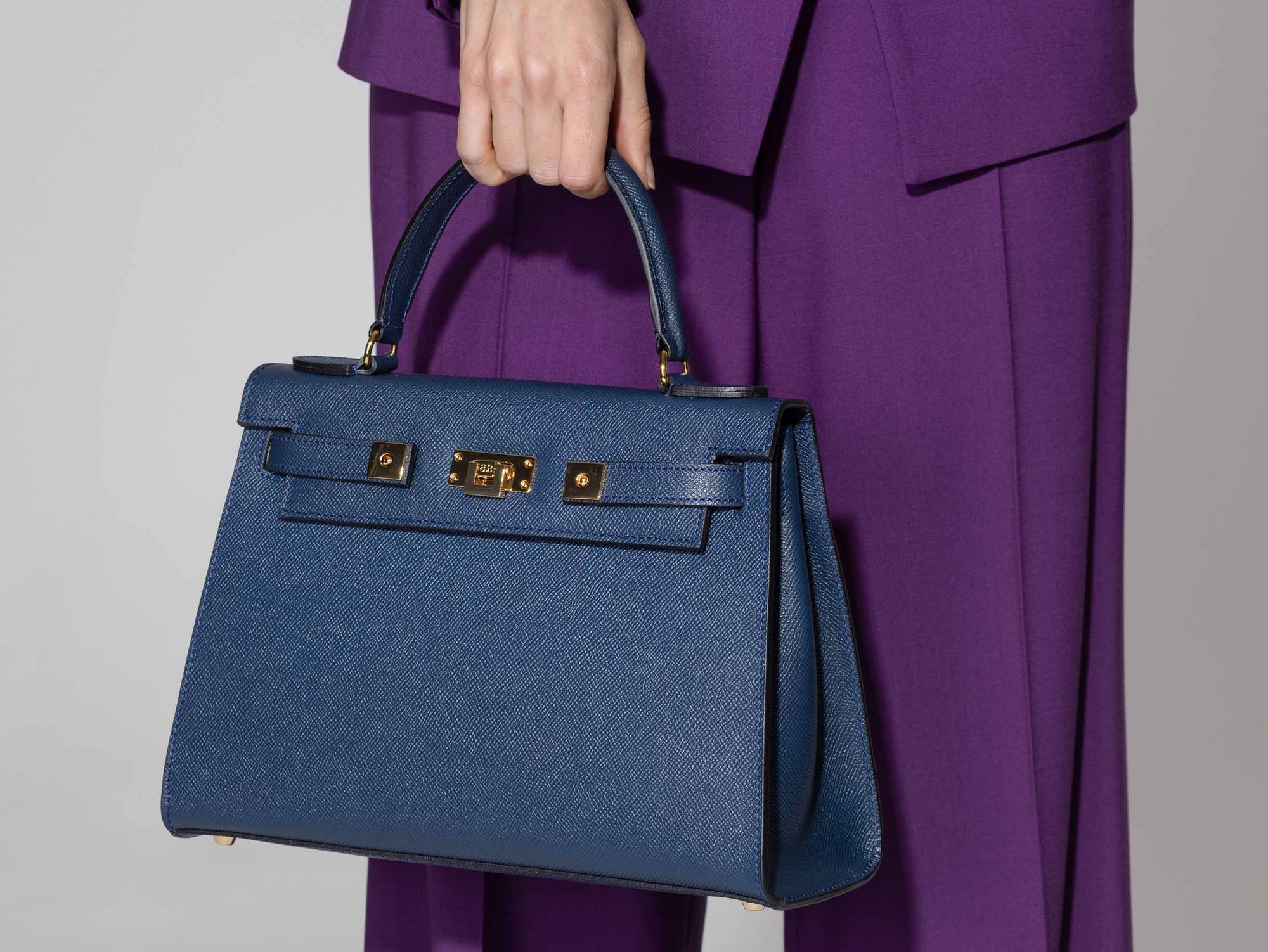 A large navy Italian leather, natural print handbag with gold hardware, a top handle with a detachable strap on a white background held by a person wearing a purple outfit on a gray background