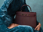 Person holding a large wine colored Italian calf leather handbag with pebble print texture, gold plated hardware, and a top handle with a detachable strap against a textured blue background