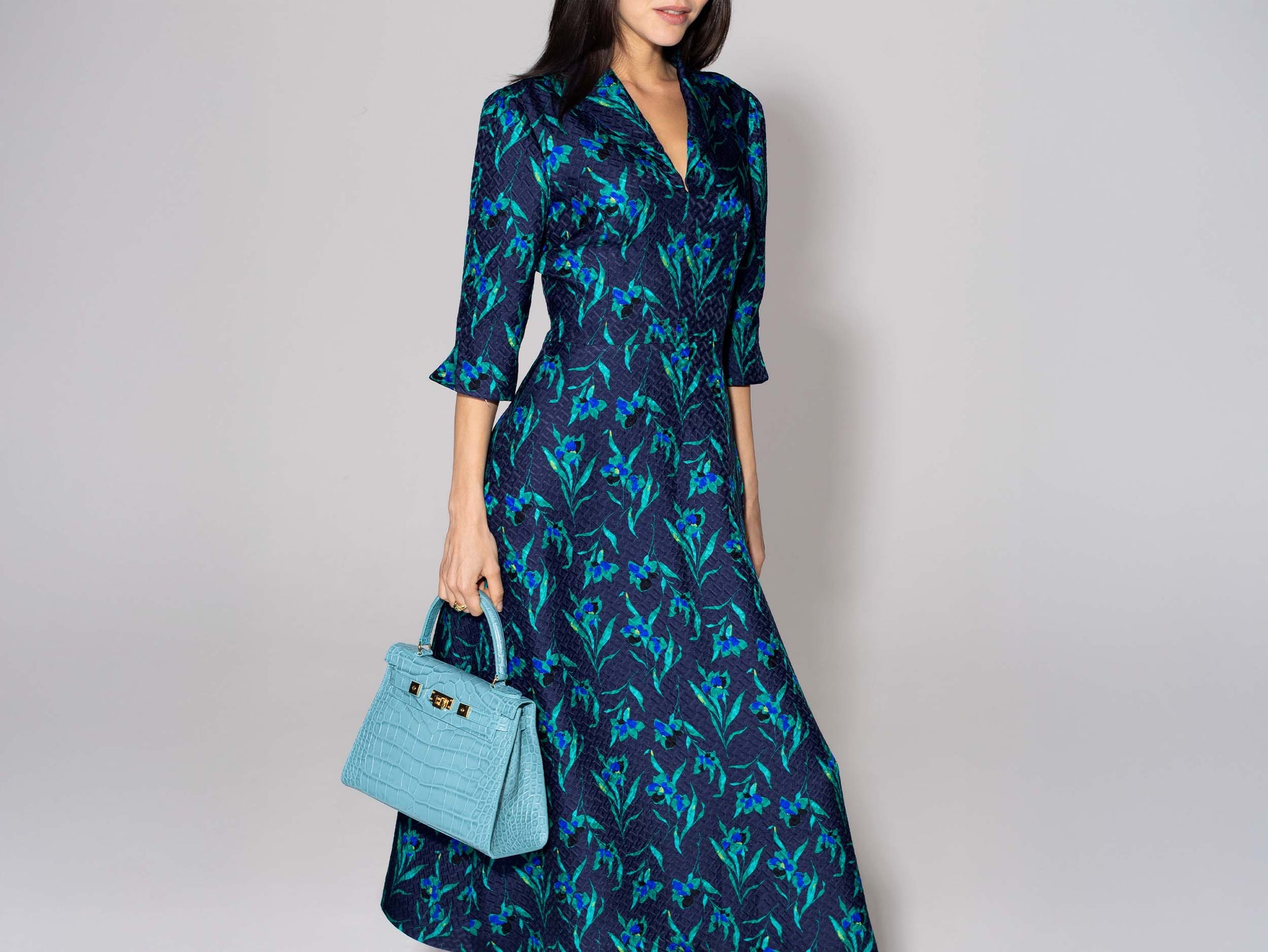 Woman wearing a blue floral dress holding a large turquoise , ‘croc’ print Italian calf leather handbag with top handle, detachable strap and gold plated hardware against a plain background