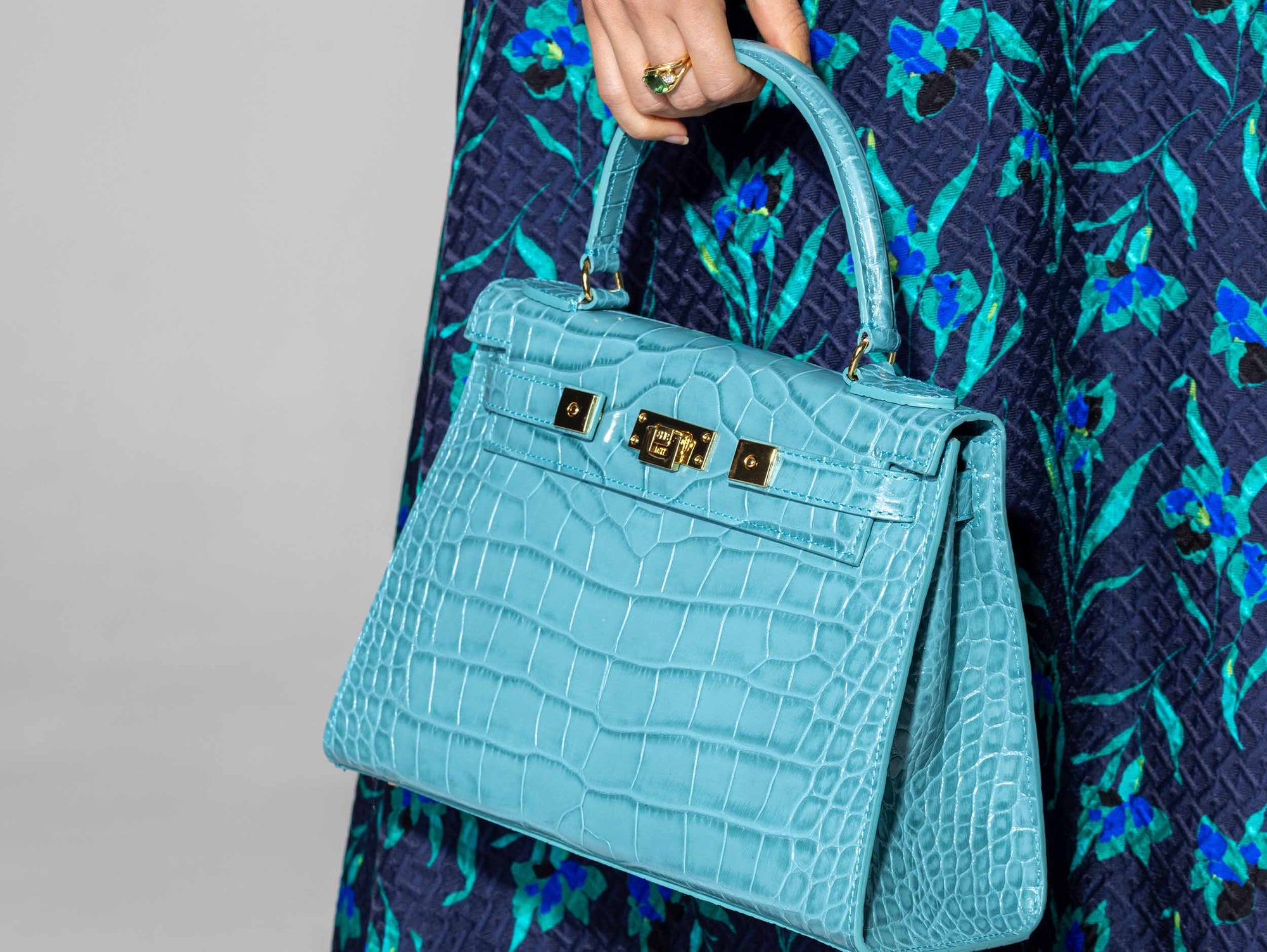 A large turquoise , ‘croc’ print Italian calf leather handbag with top handle, detachable strap and gold plated hardware held by a person wearing a dark blue floral dress.