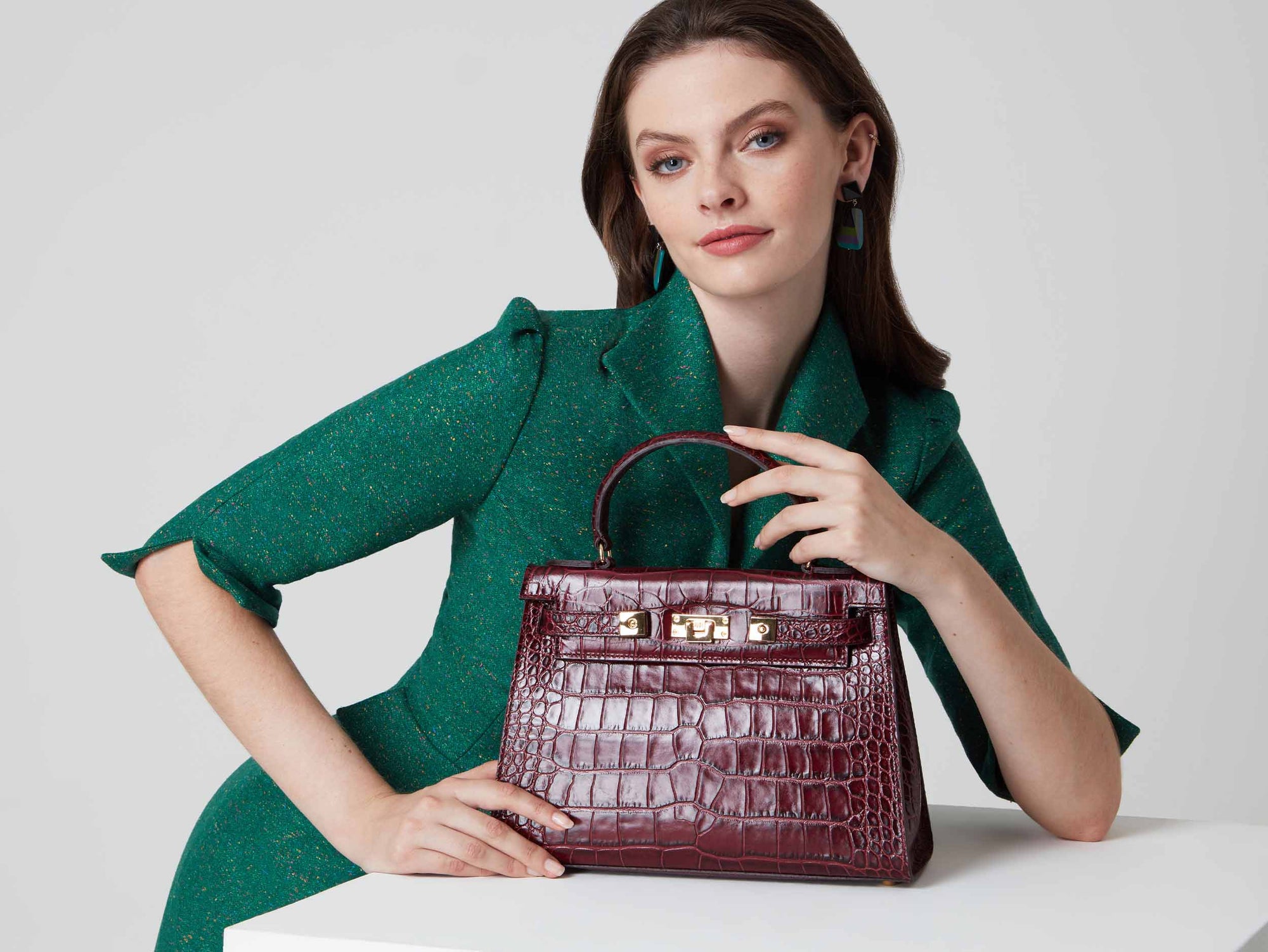 Woman holding a large wine ‘croc’ print Italian calf leather handbag with top handle, detachable strap and gold plated hardware with magnetic fastening against a plain background