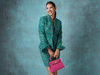 Woman in a green textured outfit holding a bright pink, medium-sized, grainy print Italian calf leather handbag with gold-plated hardware, detachable strap and a top handle against a textured blue background
