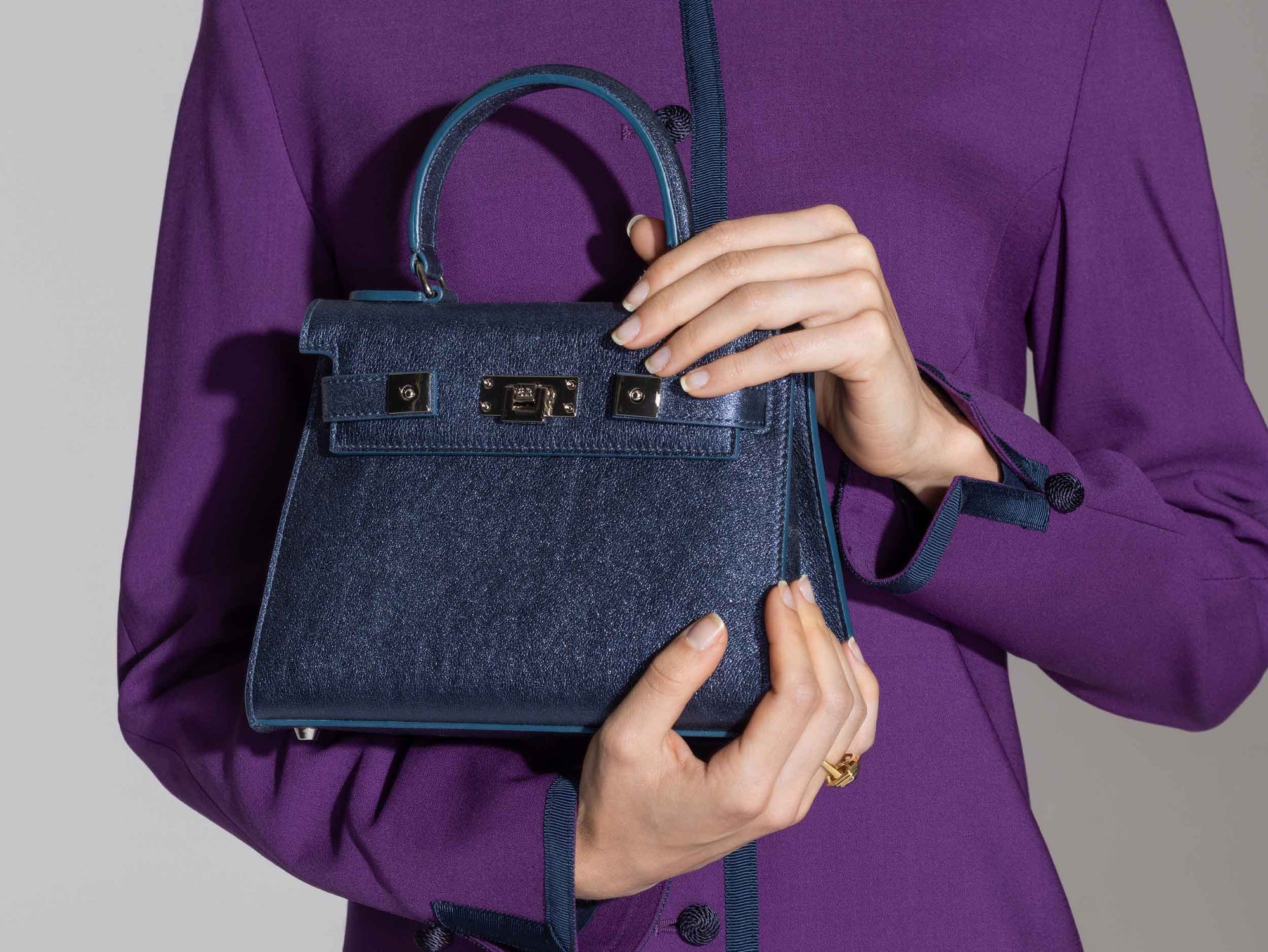 Woman holding a Medium navy blue natural grain Italian leather handbag with a hint of silver sparkle  with silver hardware, a top handle and a detachable strap with a purple coat against a gray background
