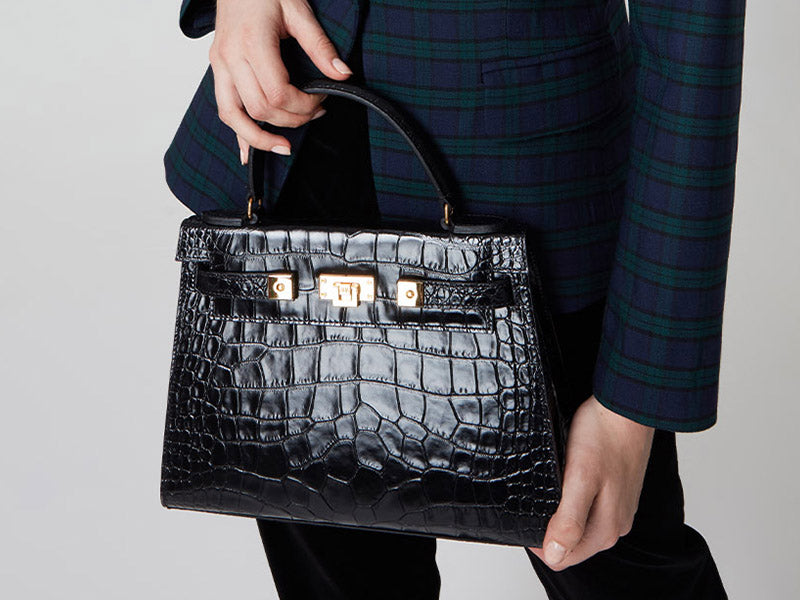 Woman holding a large black, ‘croc’ print Italian calf leather handbag with top handle, detachable strap and gold plated hardware.